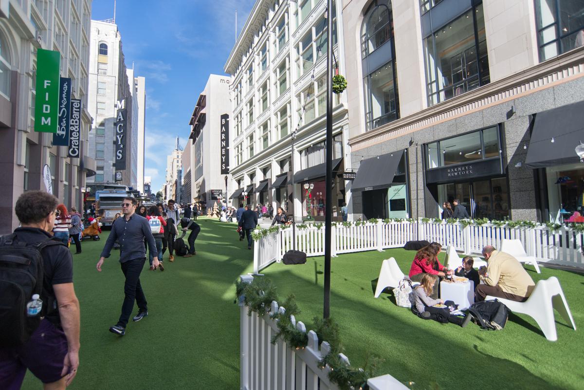 Pedestrians and visitors enjoy green space with white seating on a temporary urban plaza along a downtown street lined with buildings and storefronts.