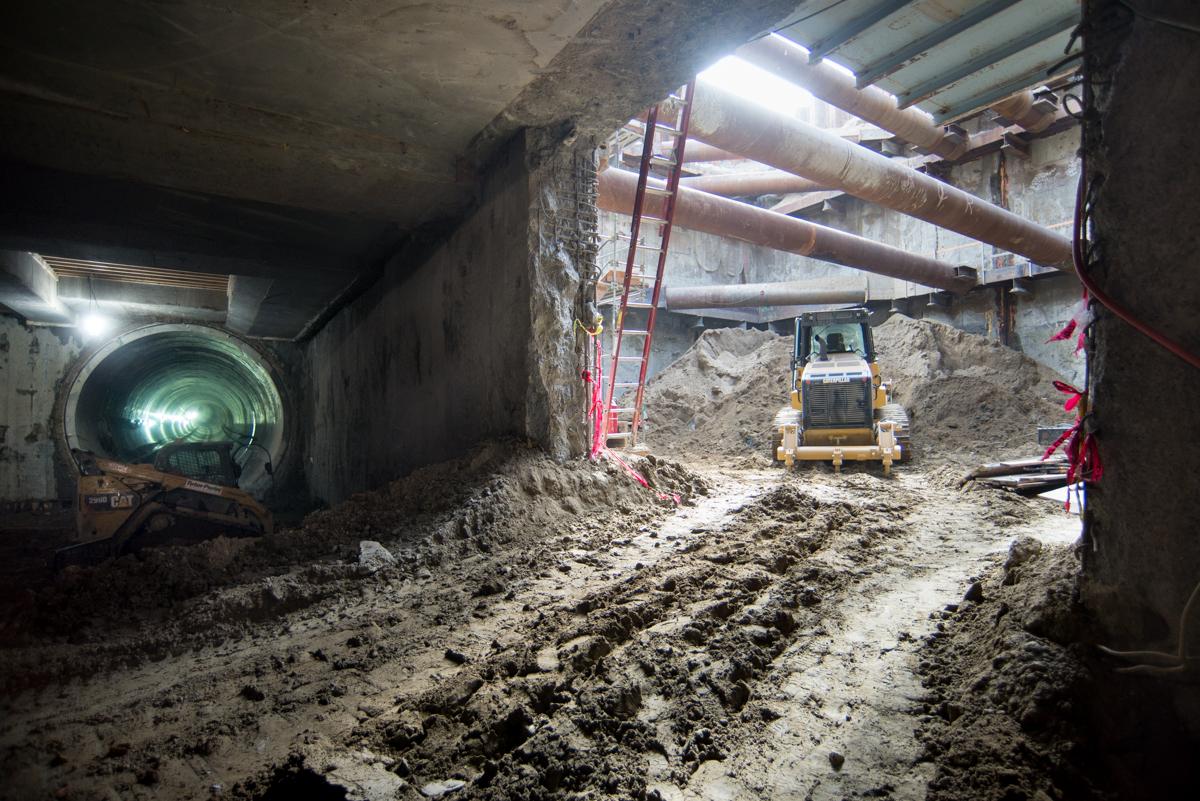Underground tunnel excavation with construction equipment, reinforcing steel bundles, and soil removal at transit station platform level.