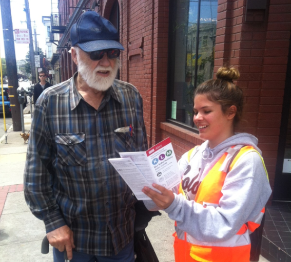 Community ambassador in safety vest assists older man with transit rider guide on city street.