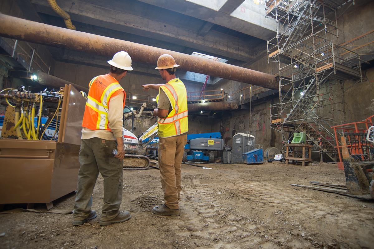 Two construction workers in safety vests and hard hats confer at an underground transit station construction site.