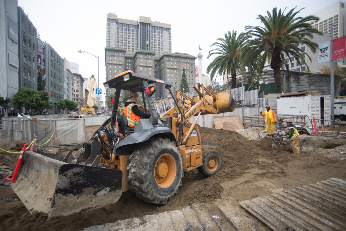 Construction crew operates heavy equipment to excavate soil and move materials on an urban street.