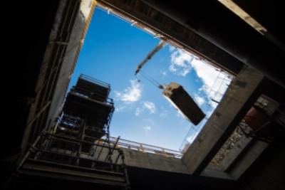 Steel bucket suspended above station headhouse excavation, viewed from below against blue sky.