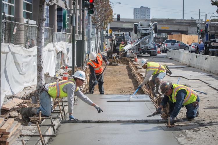 Construction workers in safety vests pouring and finishing concrete on 4th Street sidewalk during daytime urban roadway repair project.