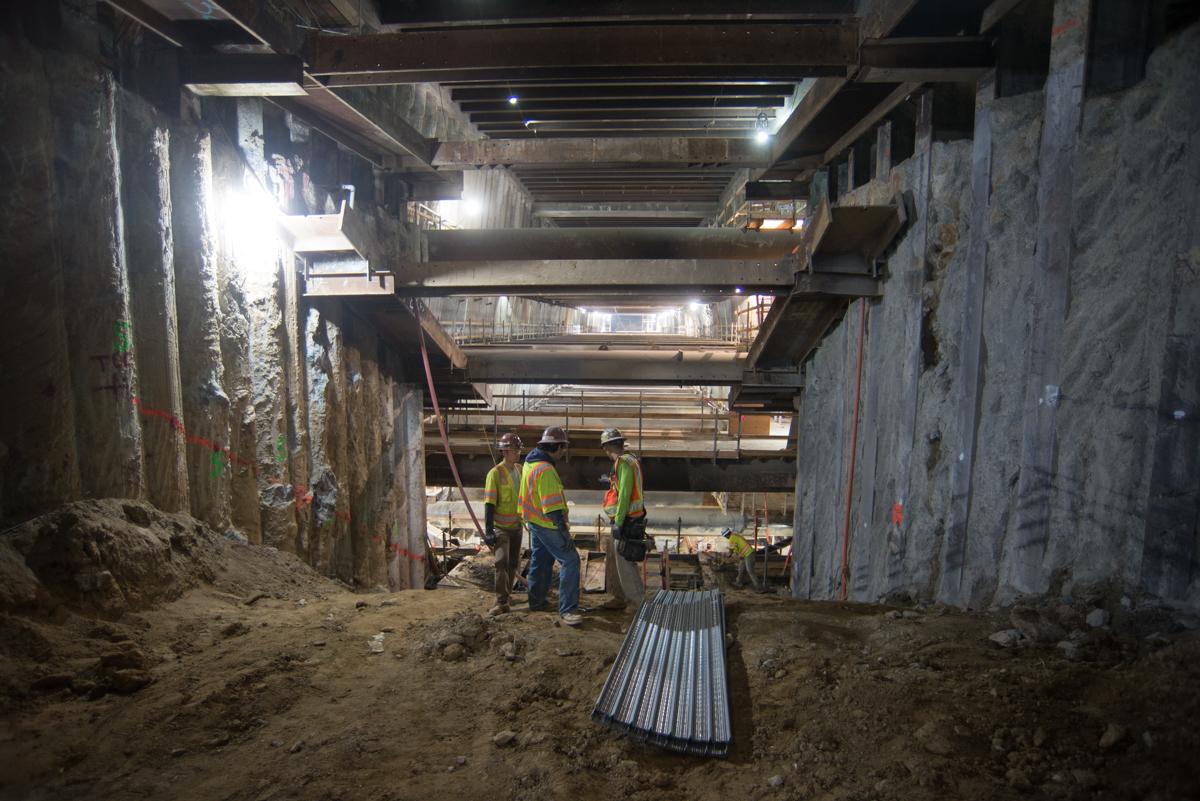 Workers in safety vests grade an incline in a concrete station box beneath highway overpass structures.