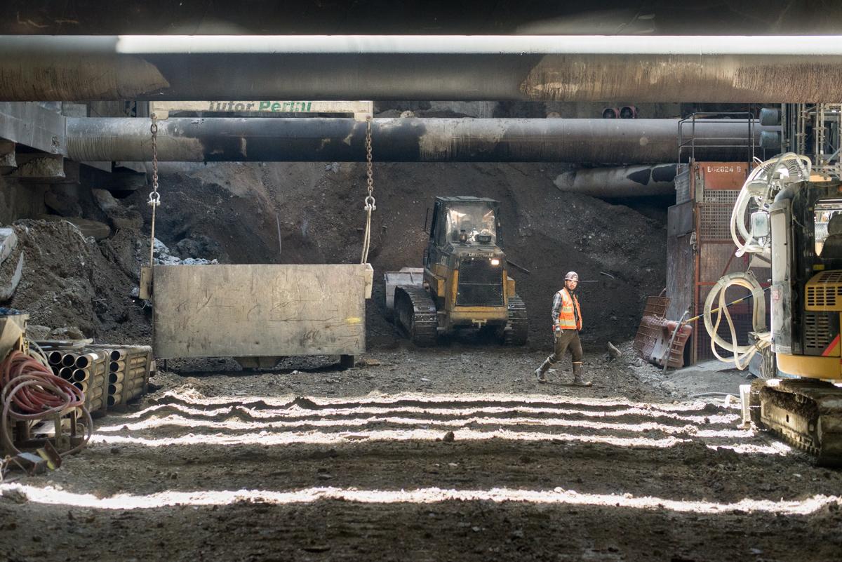 Underground construction site with excavation equipment, workers, and material being transported in steel box beneath bridge structure.