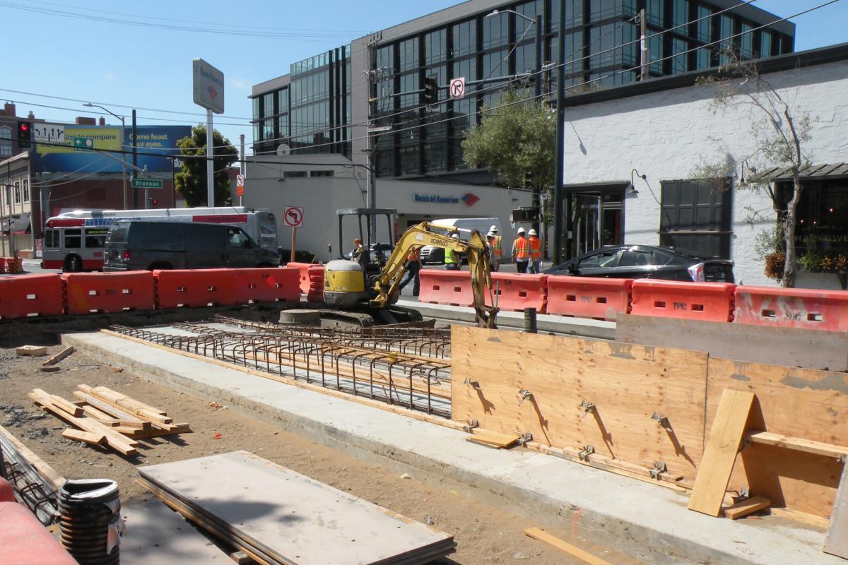 Construction site with excavator, concrete foundation, reinforced barriers, and workers near urban buildings.