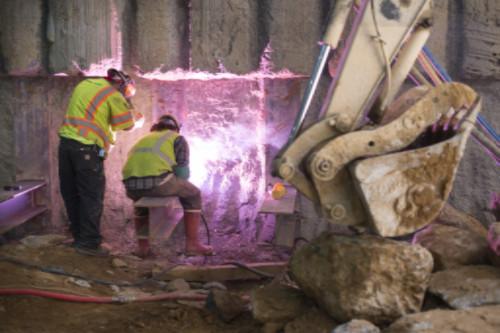 Two workers in safety vests inspect reinforced station wall at night construction site.