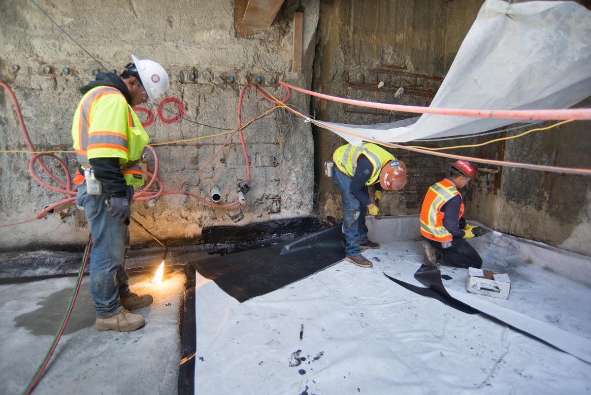 Three construction workers in safety vests installing waterproofing barrier on concrete surface with heat equipment and red cables.