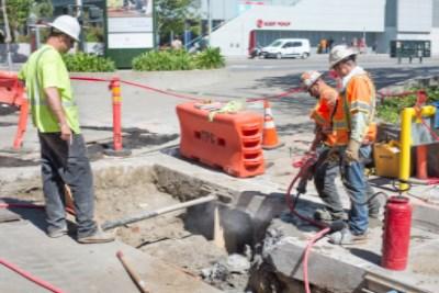 Construction workers installing water line on 4th Street near Howard Street with traffic barriers and equipment visible.