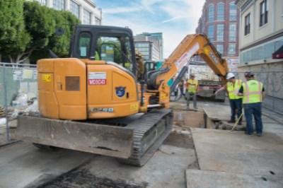 Yellow excavator performing utility work on urban street with construction workers and equipment nearby.