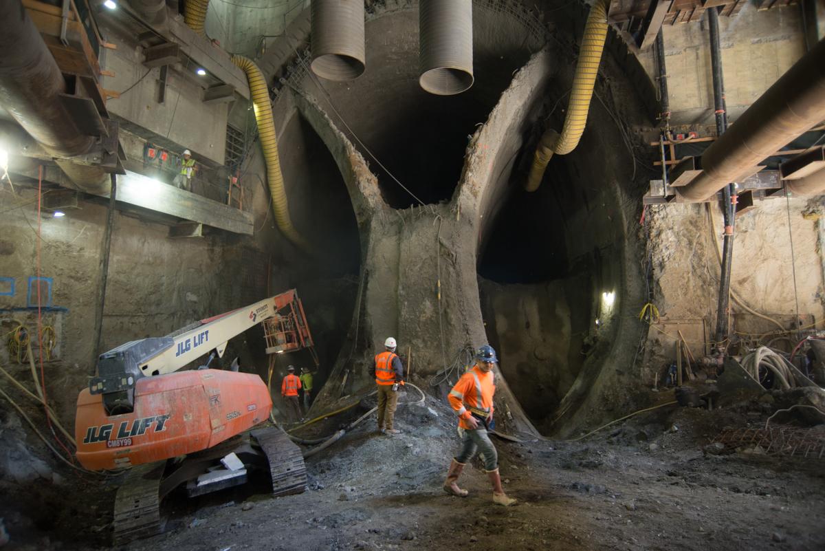 Workers in orange vests operate equipment inside a large underground cavern excavation at Chinatown Station construction site.