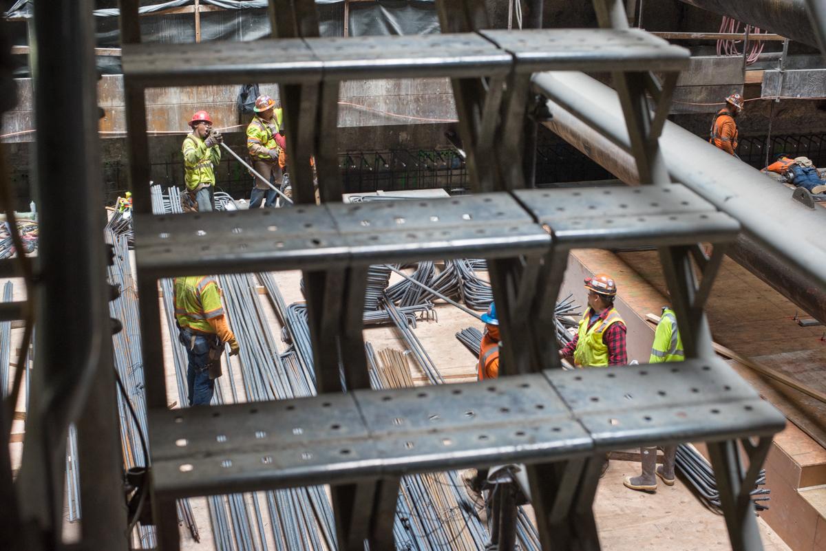 Construction workers in safety vests and hard hats work on steel framework and rebar at an underground transit station construction site.