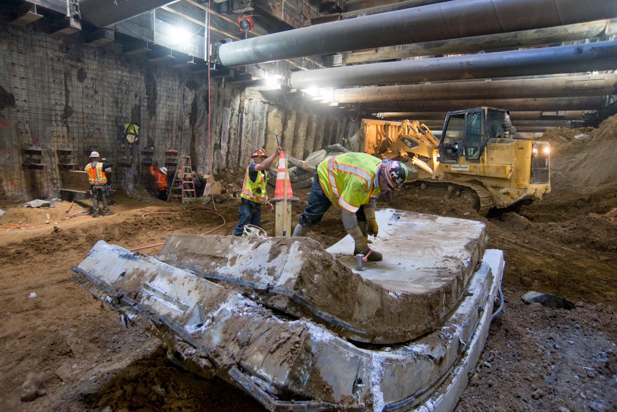 Workers remove concrete tunnel lining segments in underground station construction site with heavy equipment.