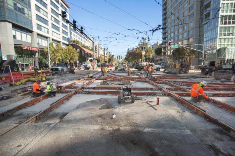 Construction workers install new streetcar track configuration on urban street with buildings and overhead lines visible.