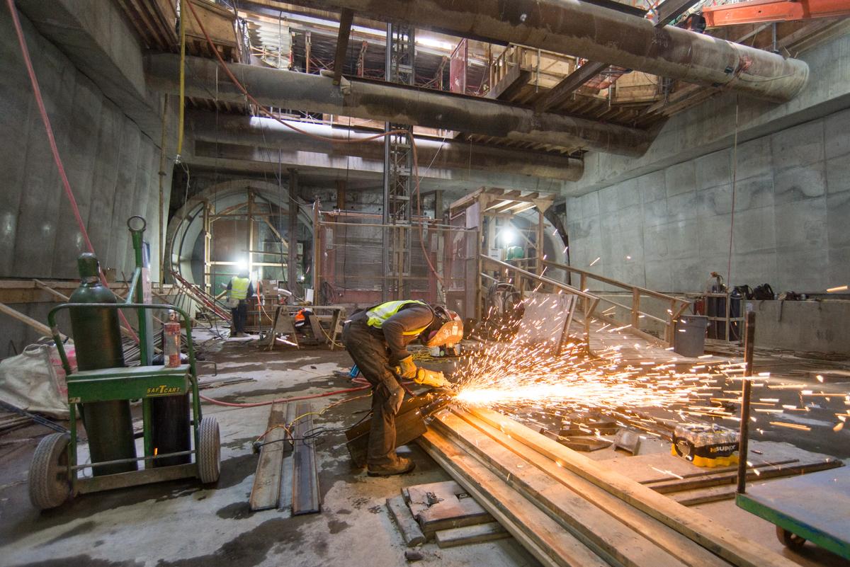 Worker welding steel bracket in underground transit tunnel construction area.