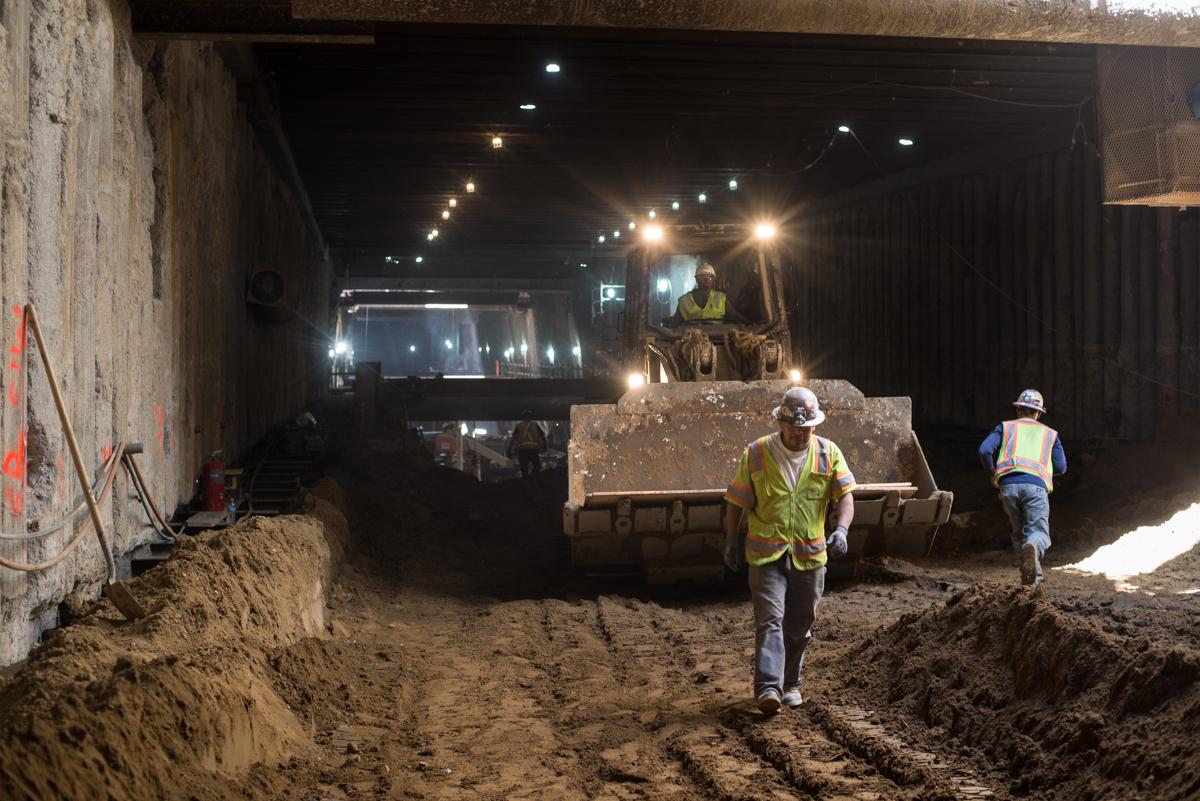 Workers in safety vests operate heavy equipment inside an underground transit station excavation site at night.