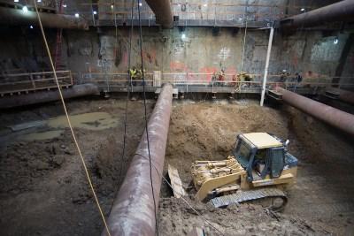 Excavation equipment removes soil from Chinatown Station headhouse construction site with steel support beams visible overhead.