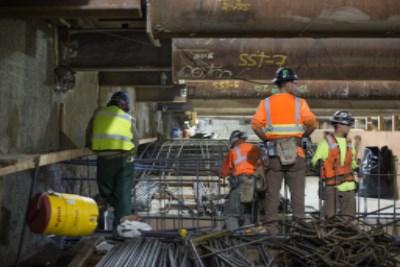 Construction workers in safety vests install underground infrastructure on an urban street.
