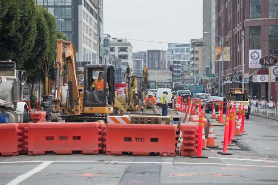 Excavator and construction equipment on 4th Street with red barriers and workers during sewer upgrades.