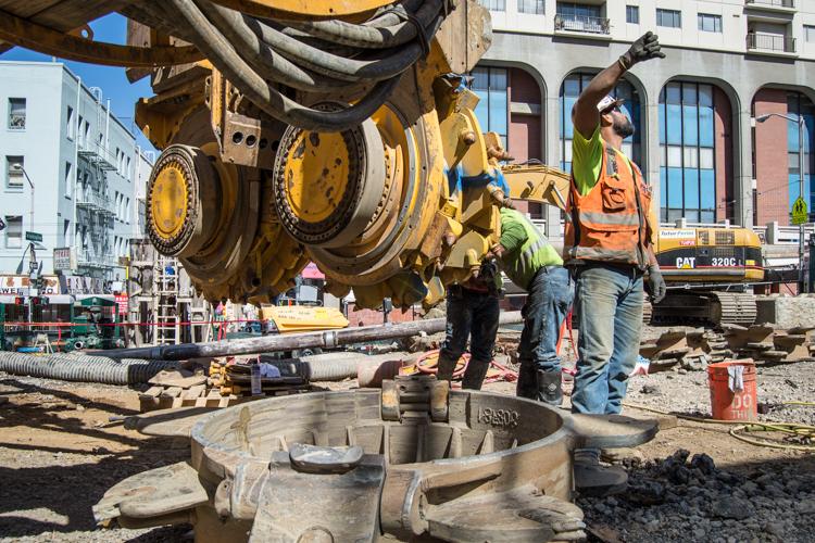 Construction crew guides crane lowering large concrete shaft component at urban transit station site.