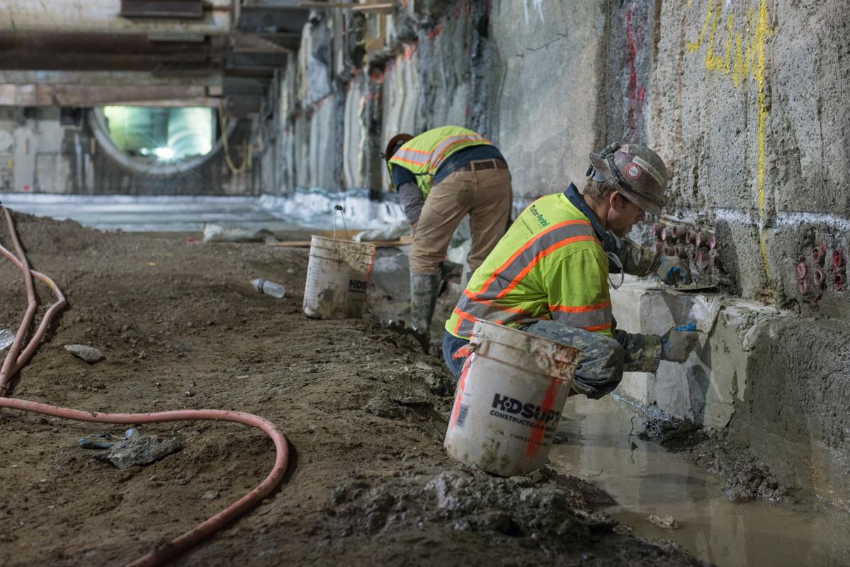Two construction workers in safety vests apply concrete to a concrete wall in an underground transit station during repair work.