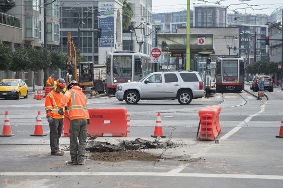Workers in orange safety vests excavate roadway at urban intersection with traffic cones and construction barriers.