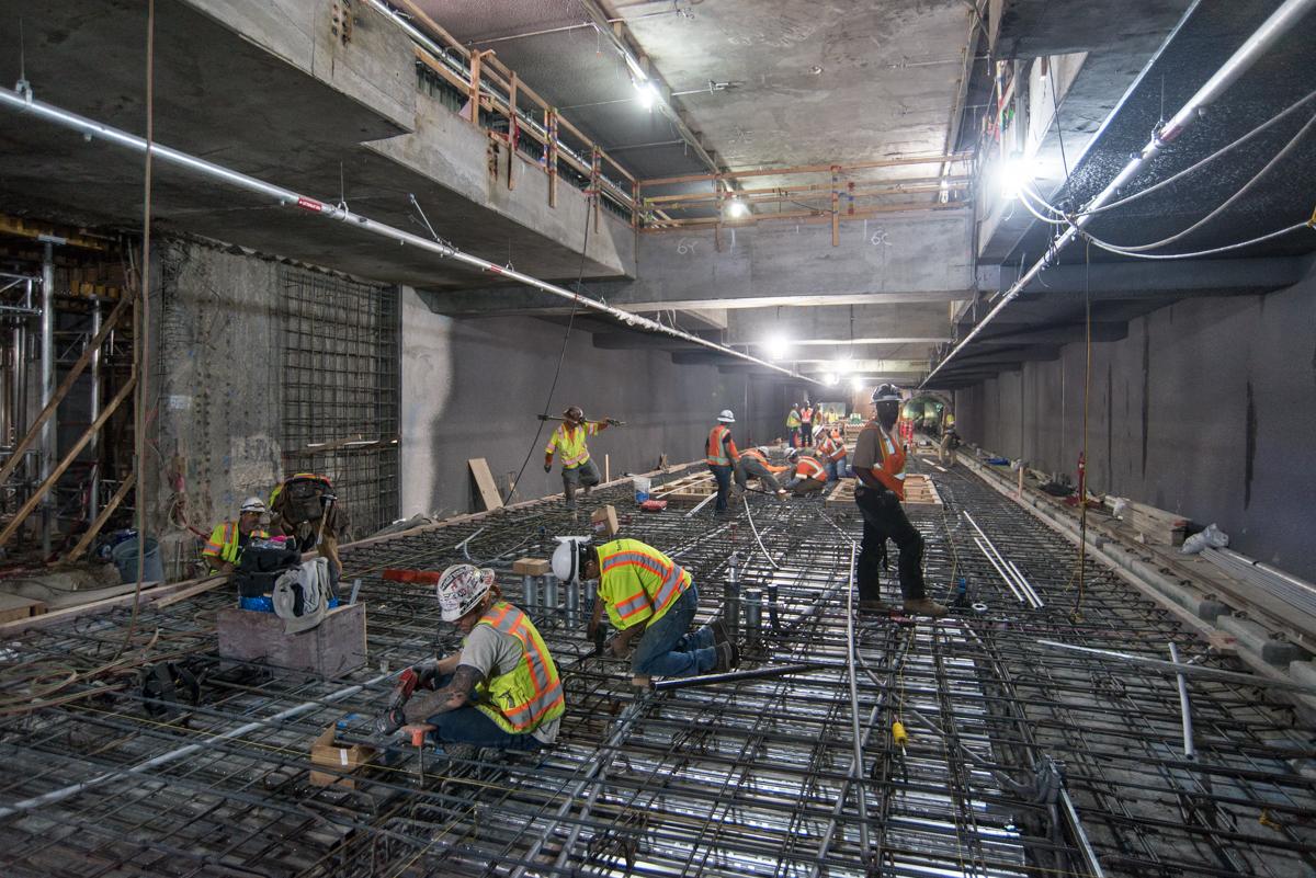 Workers in safety vests install rebar and utility conduits on concrete floor of underground transit station construction site.