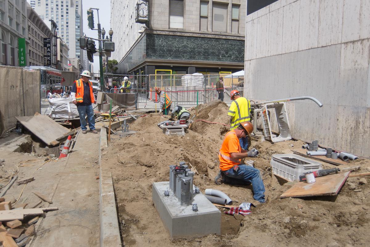 Construction workers in safety vests excavate shallow trenches for utility installation at an urban transit station site.