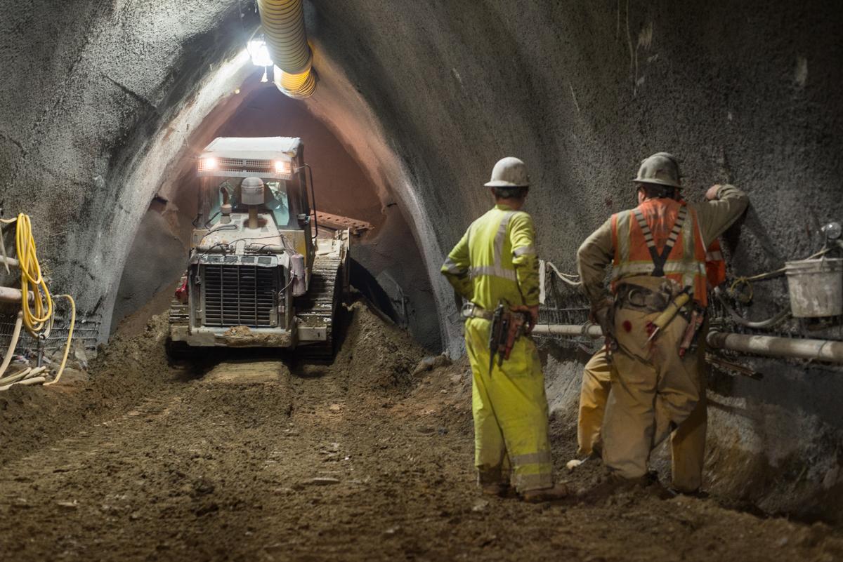 Workers in hard hats operate equipment inside an underground tunnel cavern during transit construction.
