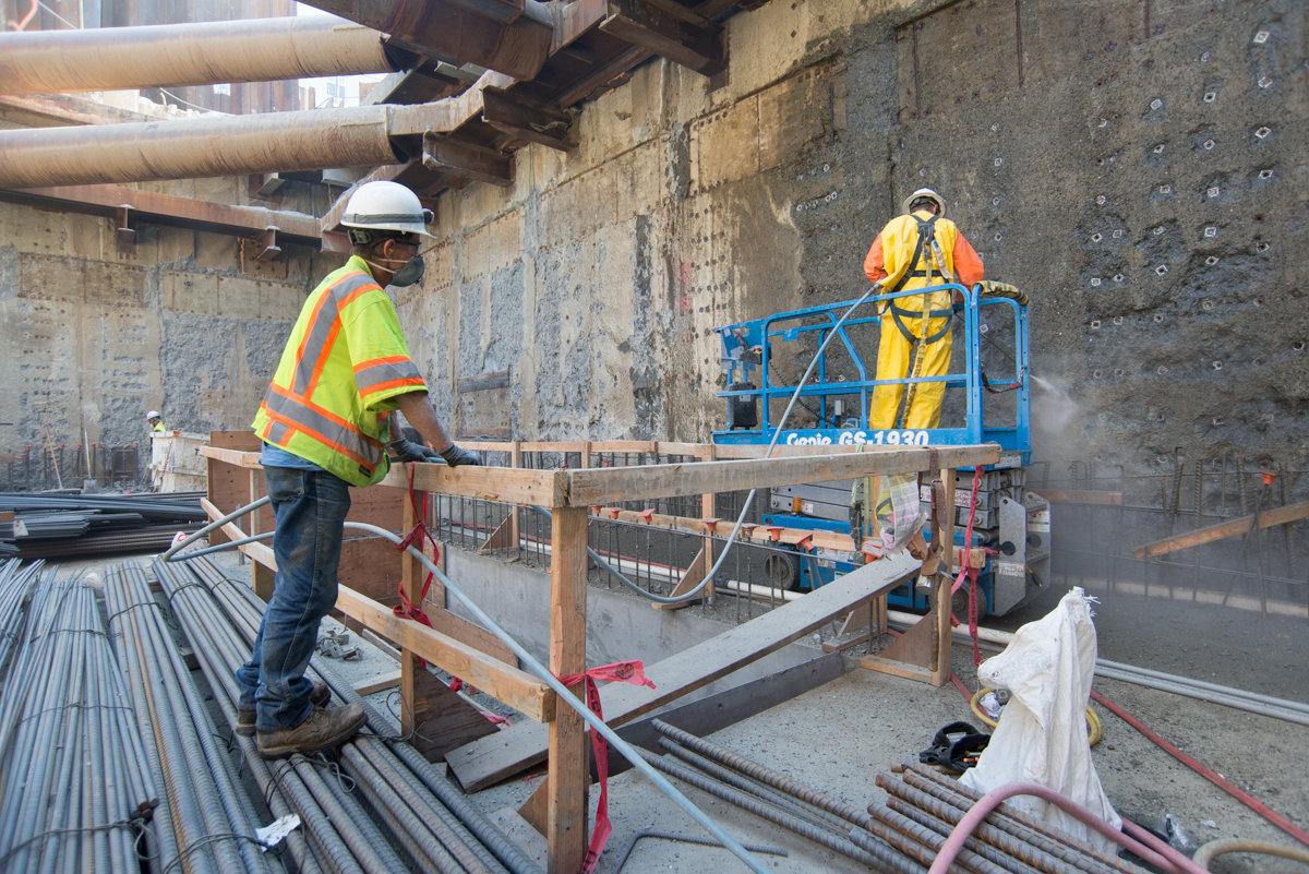 Construction workers in safety gear prepare rebar installation on concrete structure beneath elevated highway.