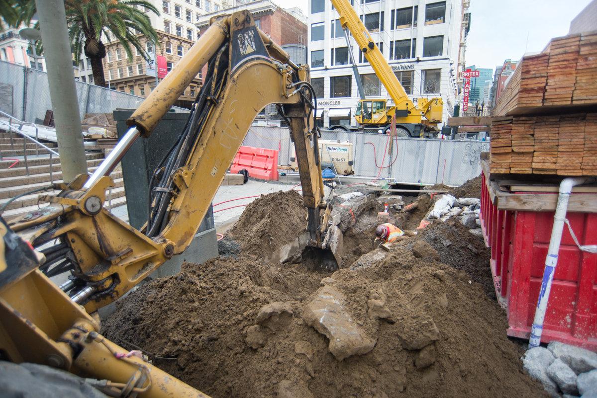 Excavator digging in soil at urban construction site with buildings and equipment visible in background.