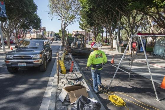 Worker in safety vest performs sewer maintenance on tree-lined street with traffic cones and equipment visible.