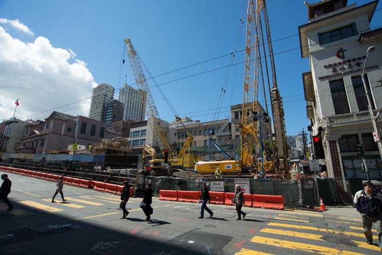 Construction site with yellow cranes and excavation equipment on Stockton Street in Chinatown, pedestrians crossing nearby.