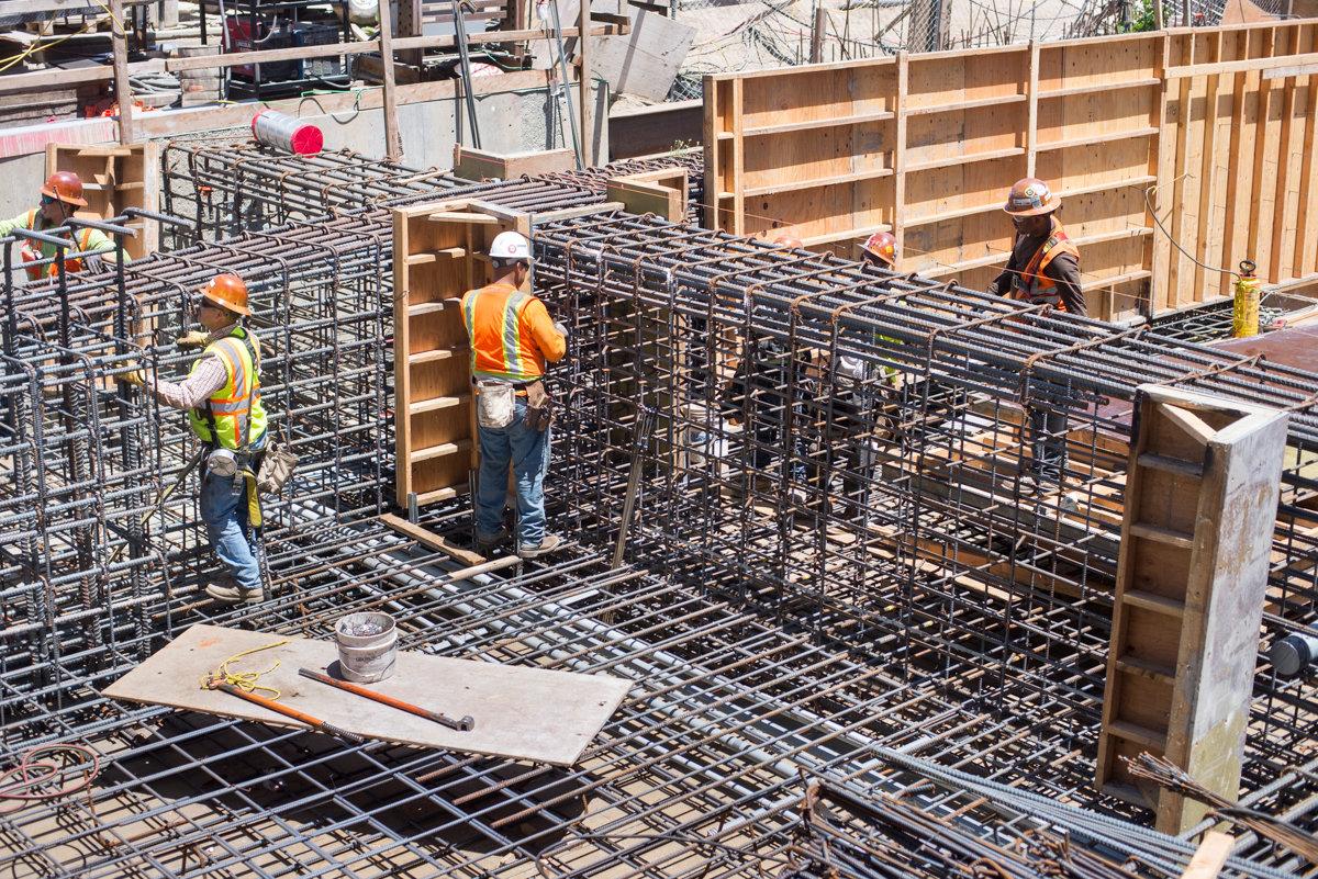 Construction workers install steel reinforcement and wooden formwork for concrete foundation at transit station headhouse.