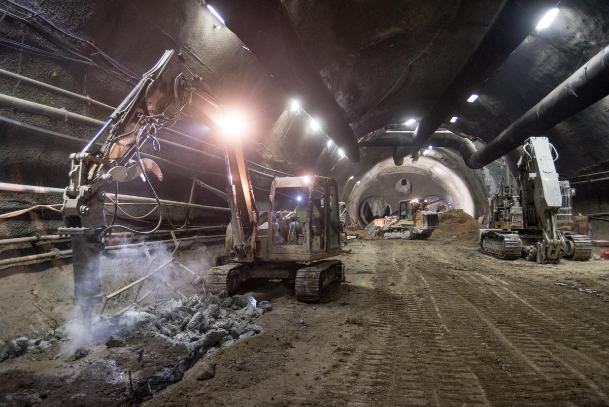 Excavator breaks apart concrete inside an underground transit station cavern at night under bright work lights.