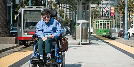 Man in wheelchair on sidewalk with Muni bus and streetcar in background