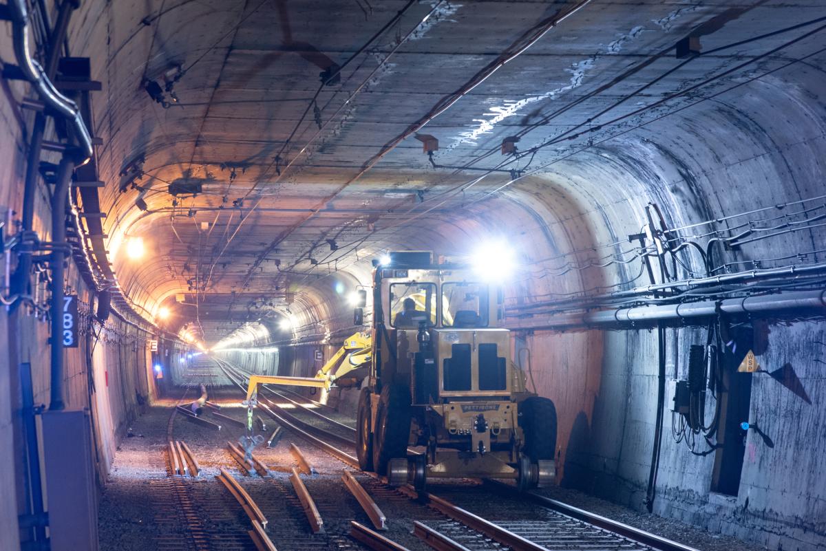Construction vehicle and workers inside illuminated subway tunnel with exposed track infrastructure.