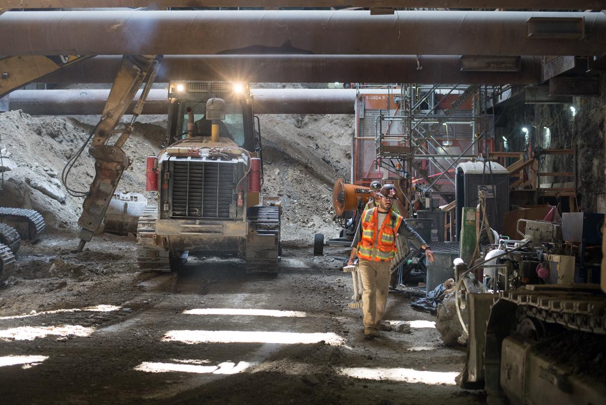 Worker in safety vest stands in underground construction site with heavy equipment and excavated material.