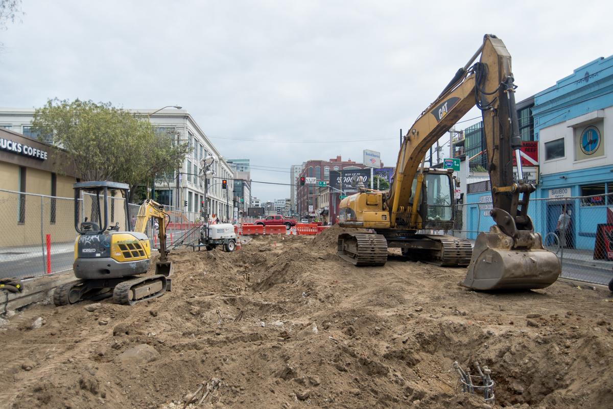 Construction site with excavators and heavy equipment on urban street surrounded by buildings.