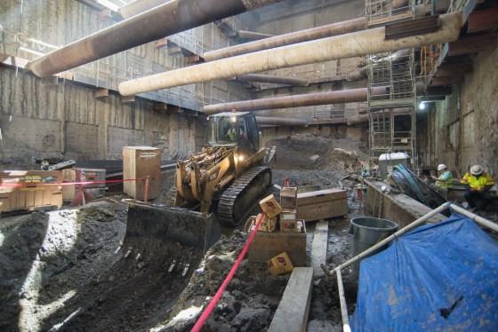 Bulldozer excavating underground station construction site with large pipes overhead and construction materials.