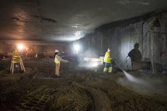 Workers in safety vests perform maintenance inside underground transit tunnel with high-pressure equipment.