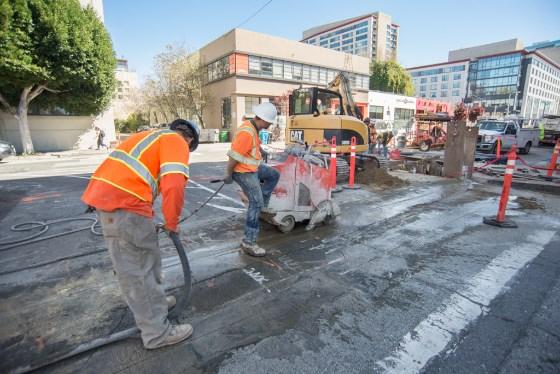 Workers in orange safety vests cut pavement with pneumatic tools during Central Subway construction on Bluxome Street.