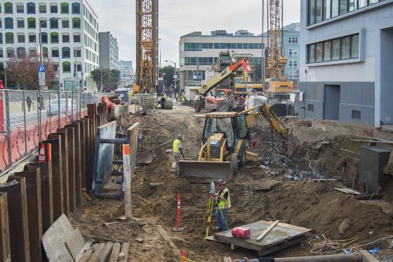 Construction excavation site on urban street with heavy machinery, workers in safety gear, and steel shoring walls.