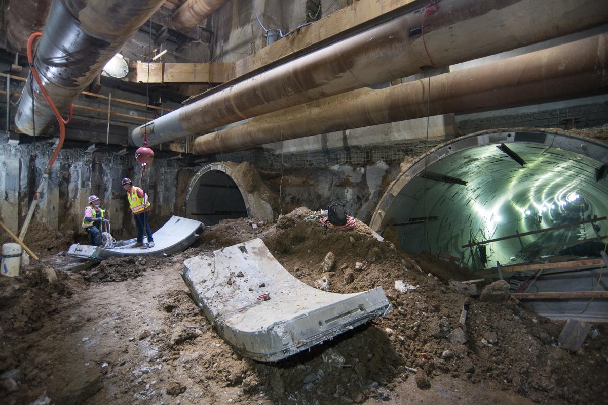 Workers in hard hats inspect underground tunnel construction site with large pipes and excavated debris.