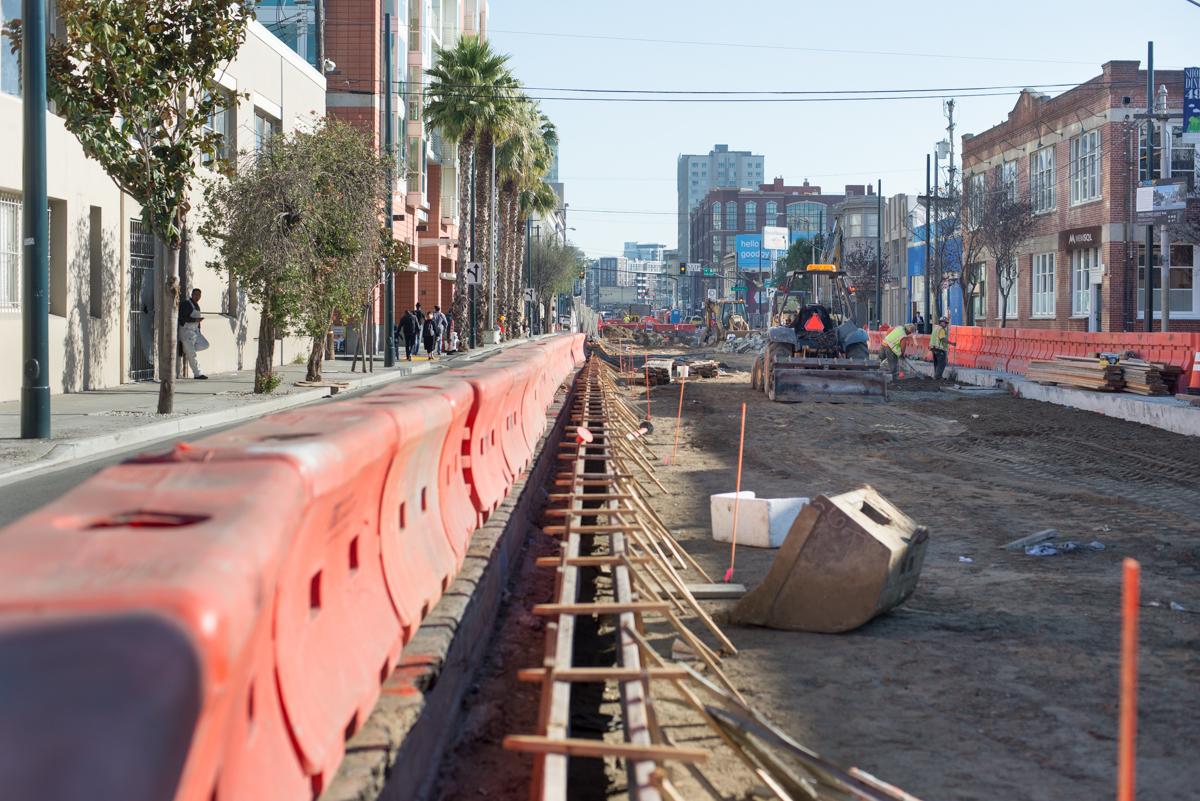 Construction workers prepare center median on 4th Street for track installation with orange barriers and concrete forms visible.