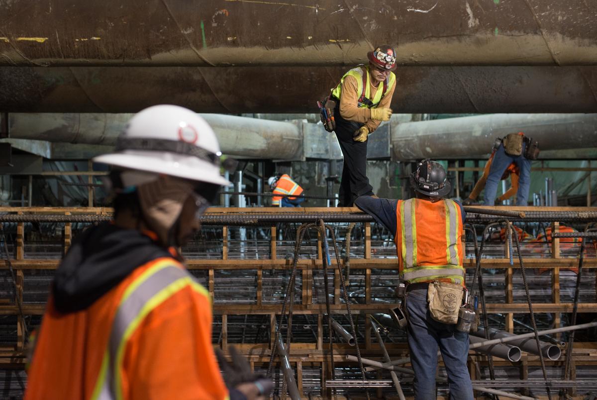 Construction workers in safety gear assemble rebar reinforcement cage at transit station platform level.