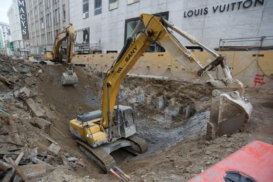 Yellow excavator removing soil and utilities at Union Square station construction site on 4th Street.