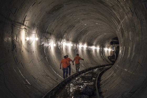 Two workers in orange vests stand in a lit subway tunnel with railroad tracks visible below.