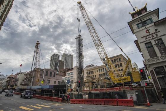 Construction cranes and equipment at Chinatown Station site with surrounding buildings and street barriers.