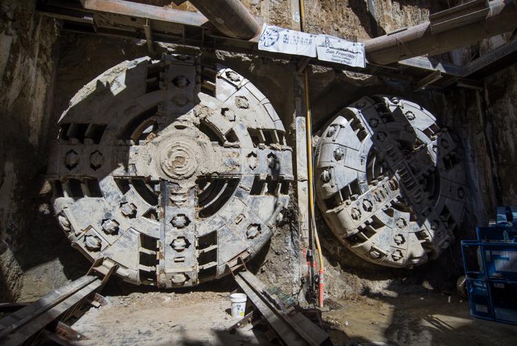 Two large circular tunnel boring machine cutterheads displayed side by side at a construction site, with concrete structures and metal framework visible above.